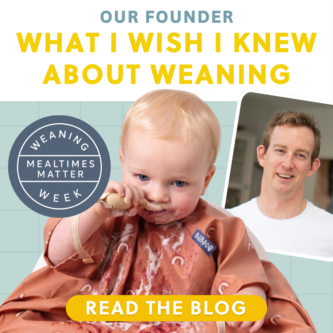 Messy baby in a terracotta bib eating with a wooden spoon, next to a smiling man.