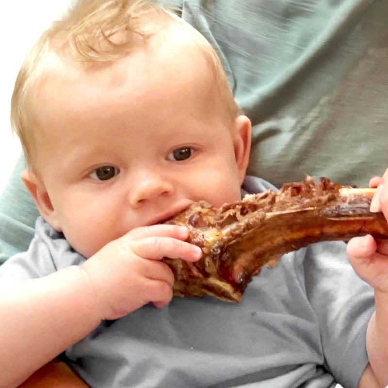 Blonde baby in a grey top gnawing on a large, dark bone, held by an adult in a green top.