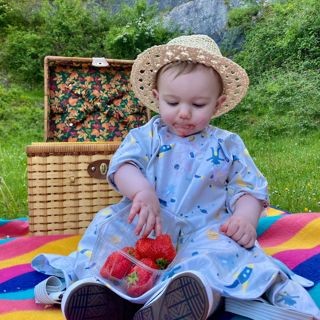 Baby in straw hat and bib, eating strawberries on a colourful picnic blanket with a wicker hamper.
