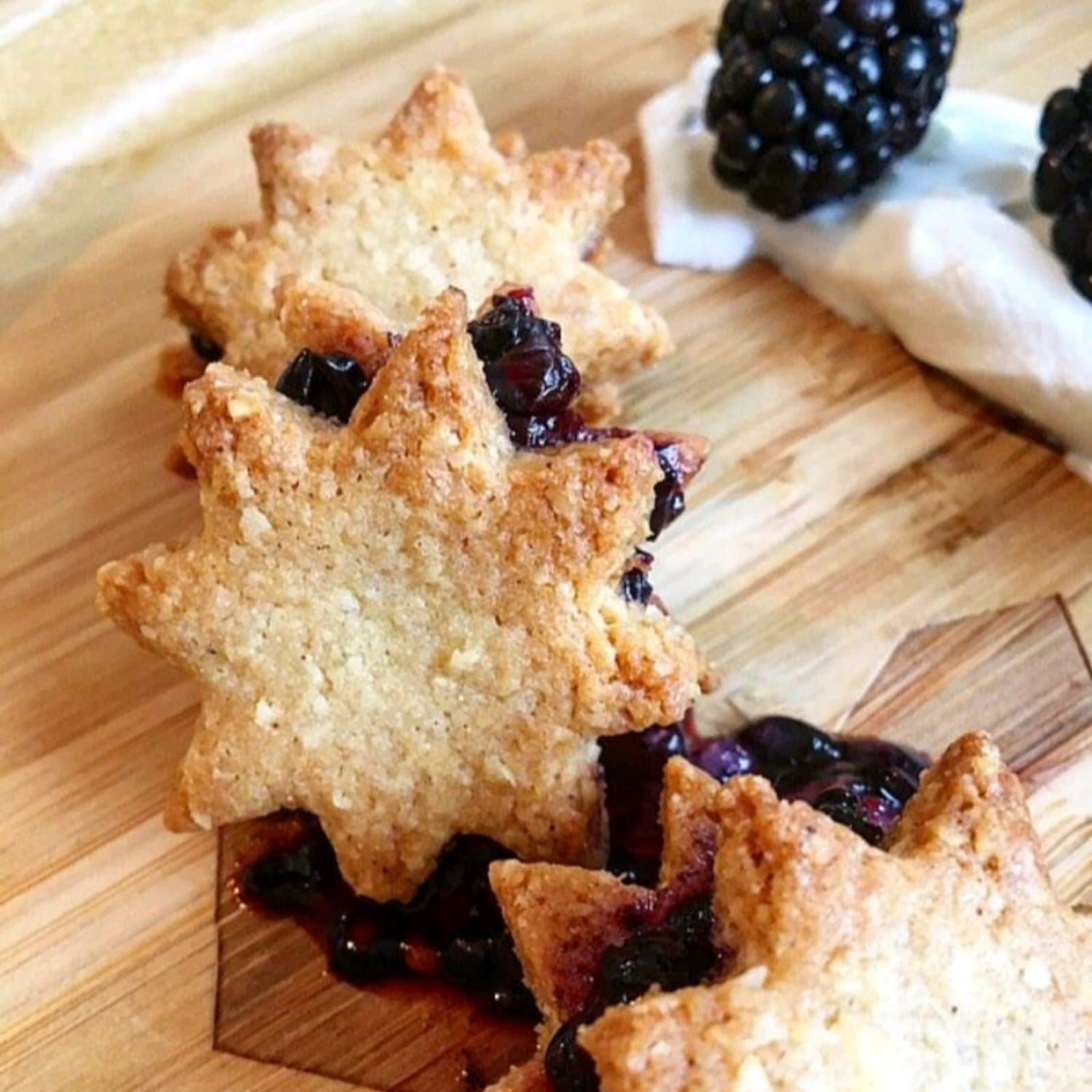 Golden star-shaped biscuits filled with dark berry jam, on a wooden board with fresh blackberries.