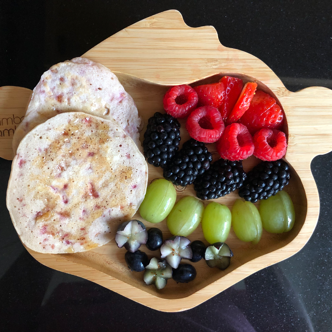 Fluffy raspberry pancakes on a bamboo plate with fresh berries and grapes.