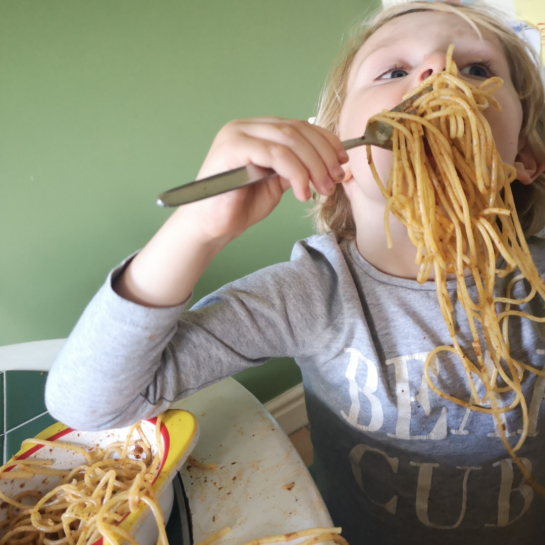 Young child with blonde hair eating a large forkful of messy spaghetti against a green wall.