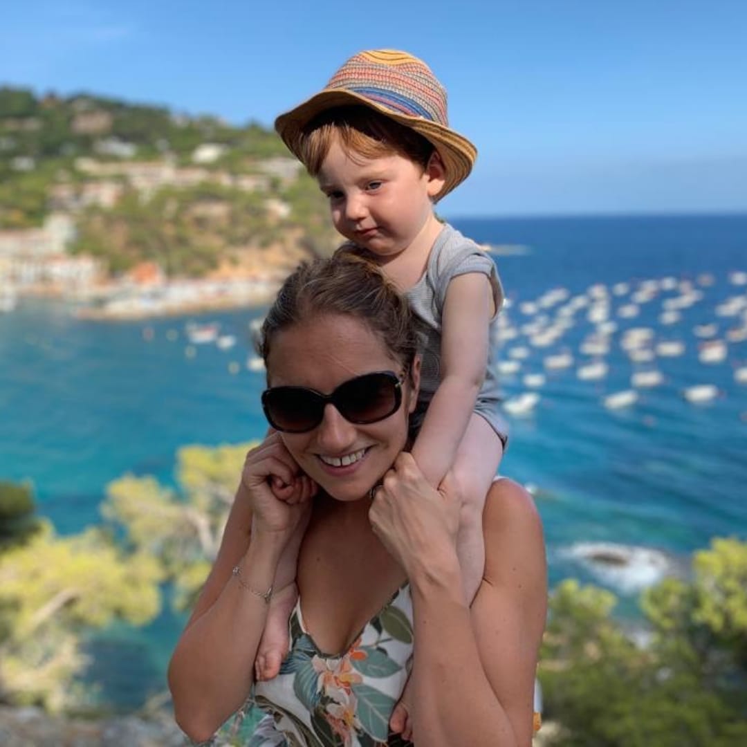 Smiling mum with sunglasses carrying her young child in a straw hat on her shoulders, overlooking a sunny blue bay.