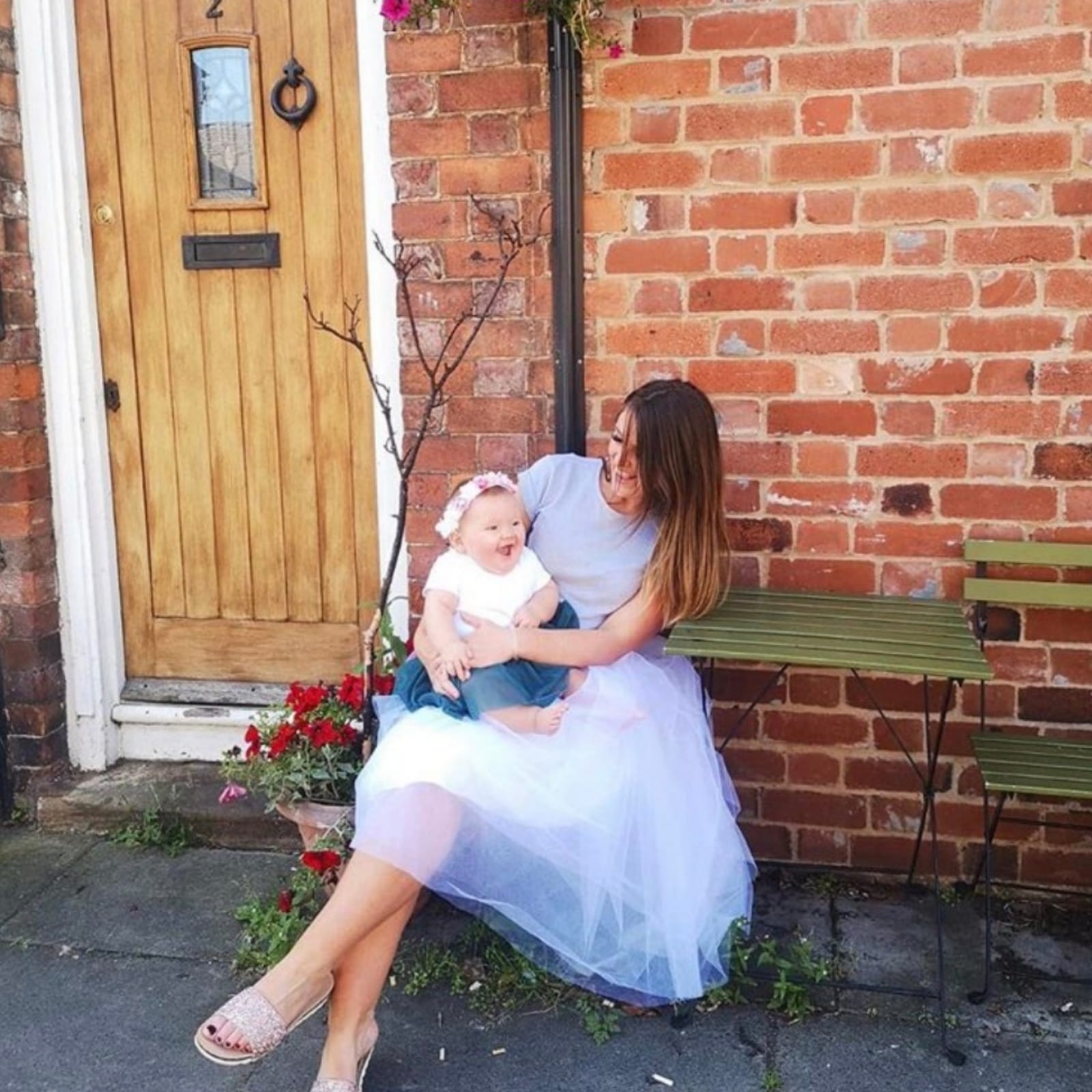 Smiling mum in a white tulle skirt holding a laughing baby, sitting outside a brick house.