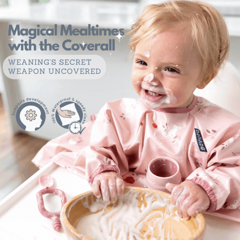 Happy baby in a pink patterned coverall bib, covered in food during weaning.