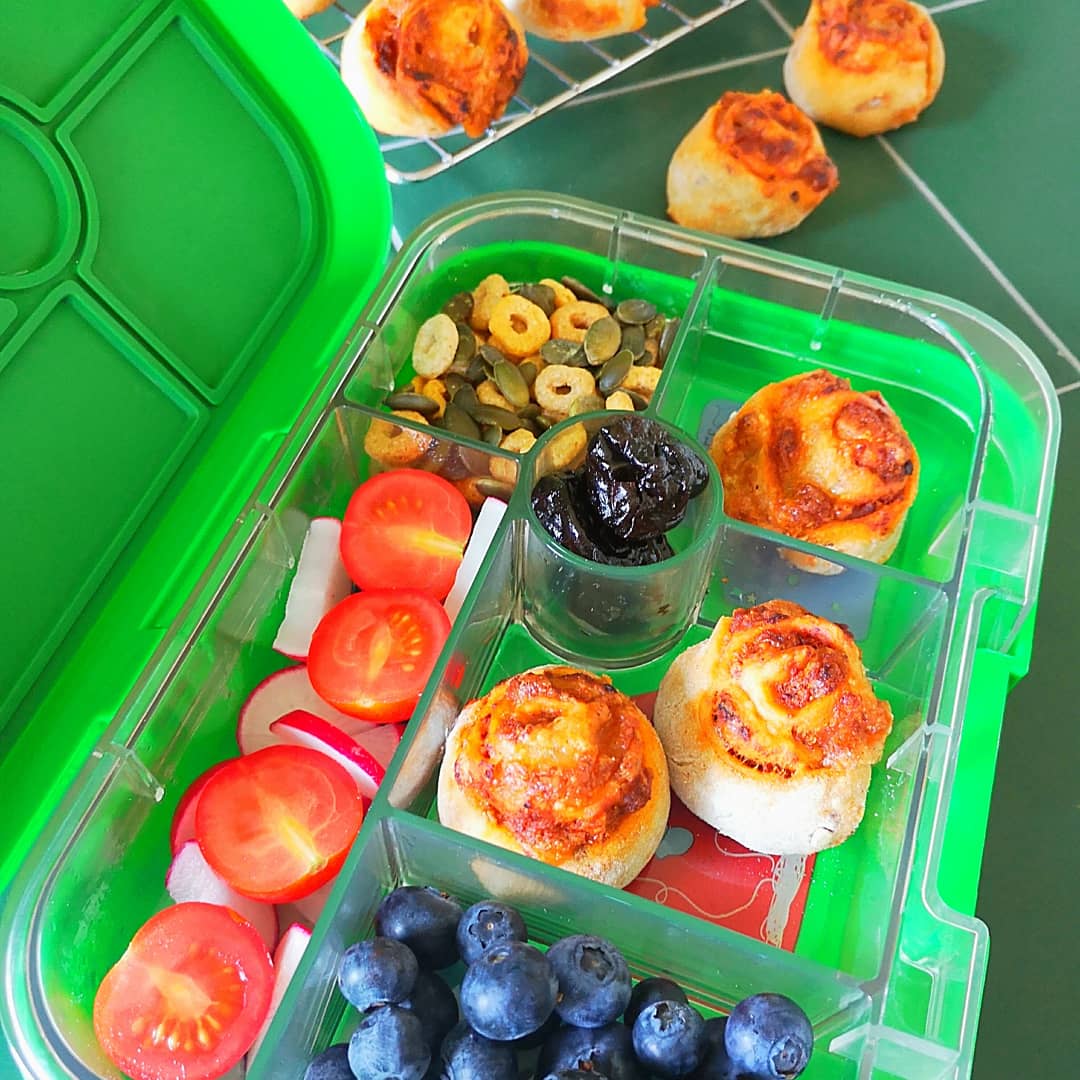 Bright green bento box filled with mini pizza splats, blueberries, cherry tomatoes, and seeds.