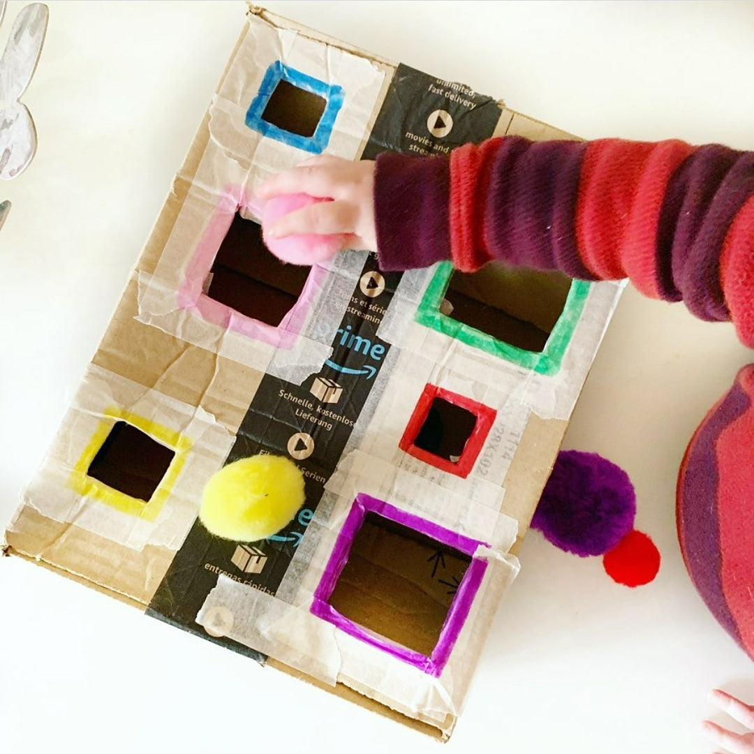 Child's hand sorting a pink pom-pom into a homemade cardboard colour sorting box with coloured squares.