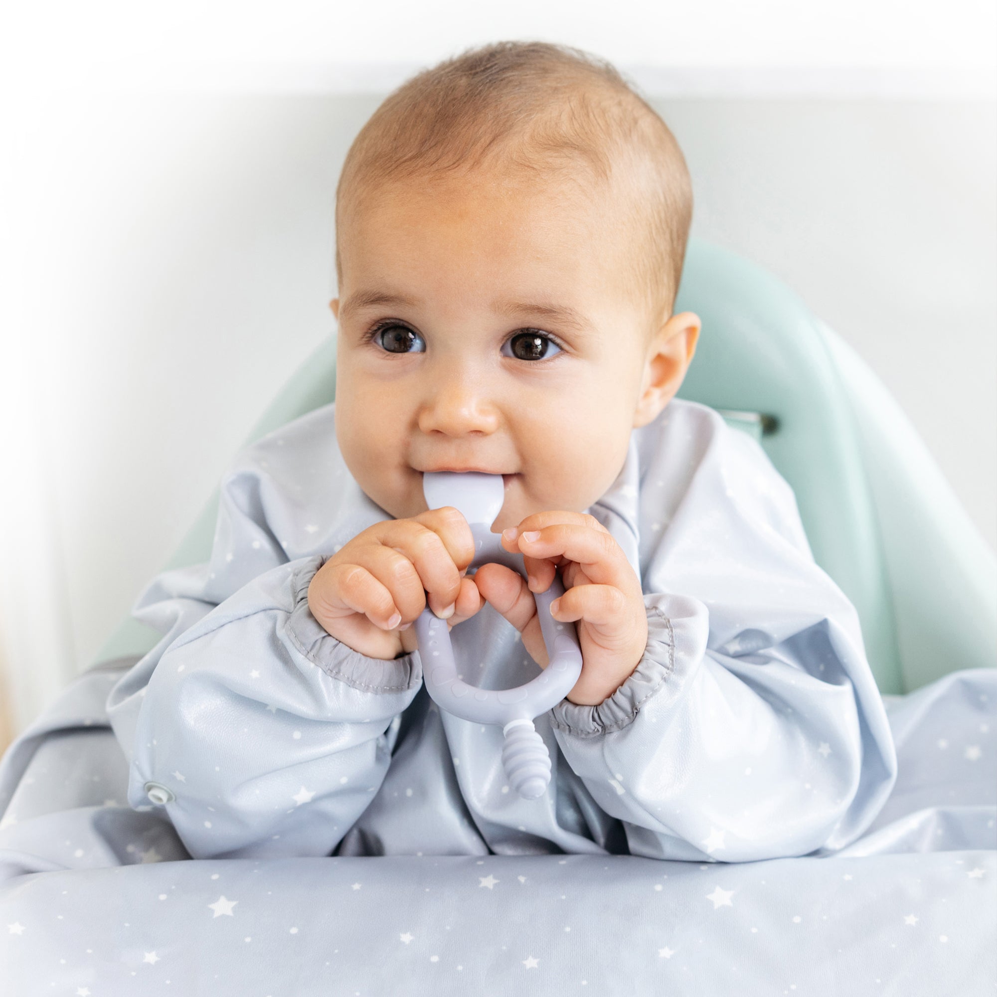 Happy baby in a star-patterned bib, holding a grey silicone weaning spoon and teether.