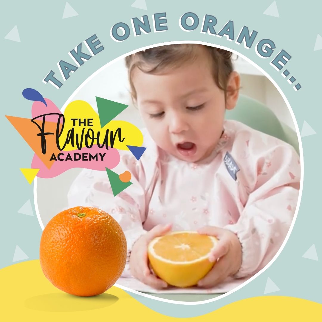 Baby in a pink bib holding a halved orange, with a whole orange beside it.