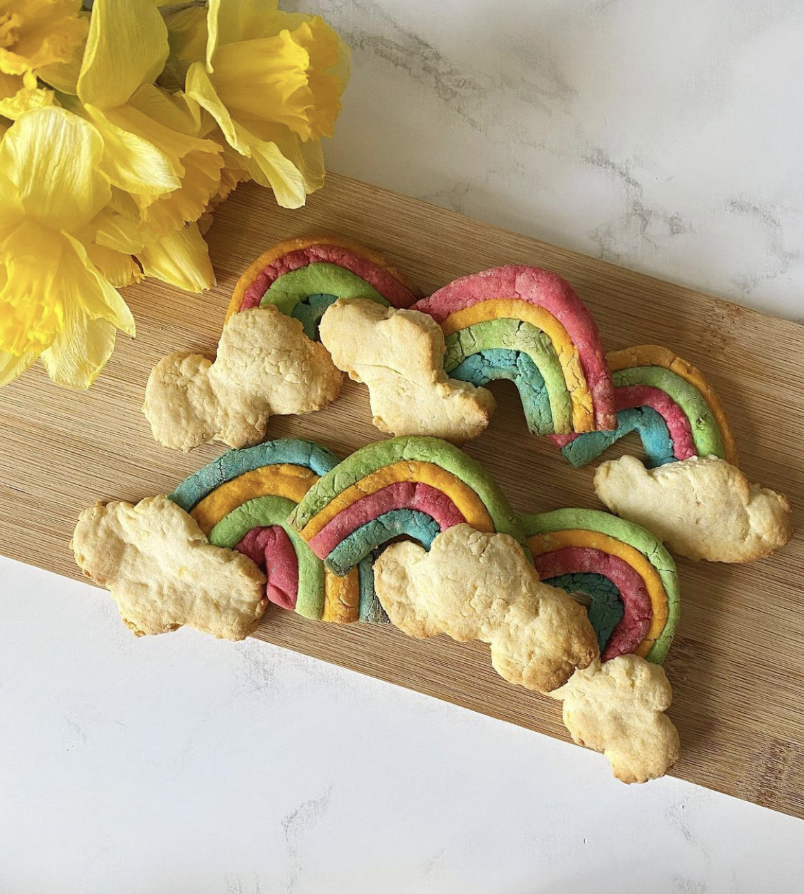 Vibrant vegan rainbow cookies with fluffy cloud ends on a wooden board, beside yellow daffodils.