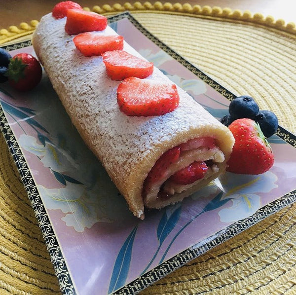 Strawberry roulade dusted with icing sugar, garnished with fresh strawberries and blueberries on a decorative plate.