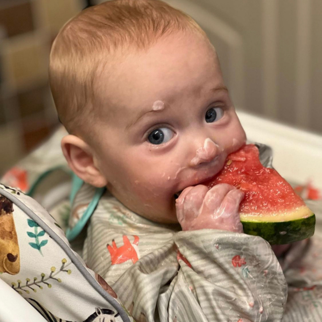 A messy baby with blue eyes eating a slice of red watermelon in a highchair.