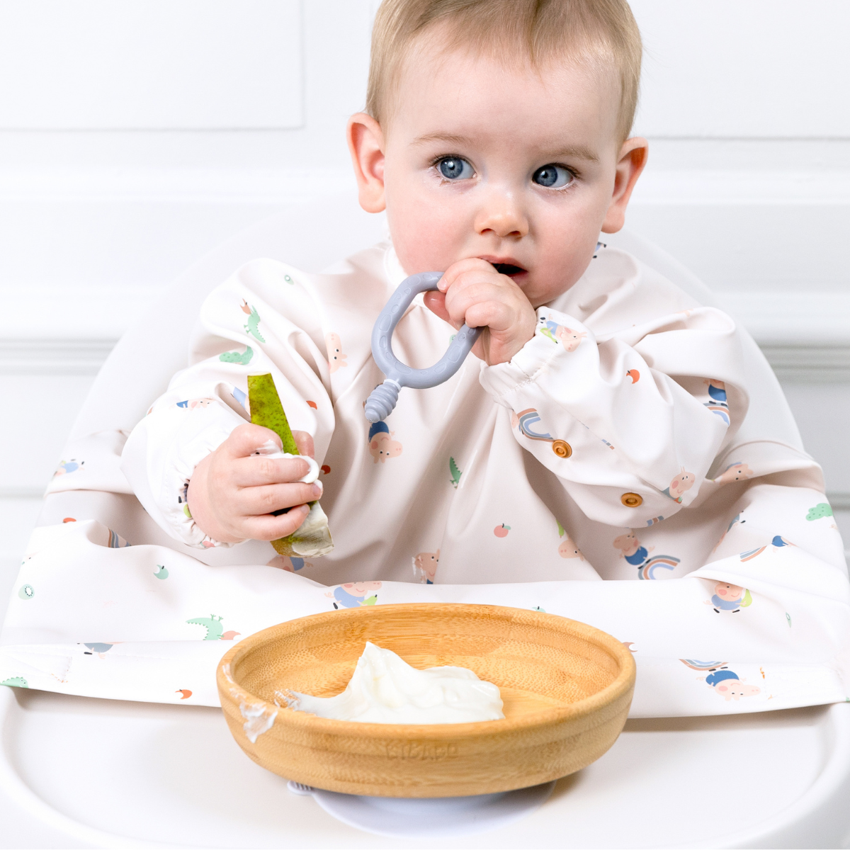 Baby in a patterned bib holding a teether and green food, with a bamboo suction bowl of yogurt. - Product image