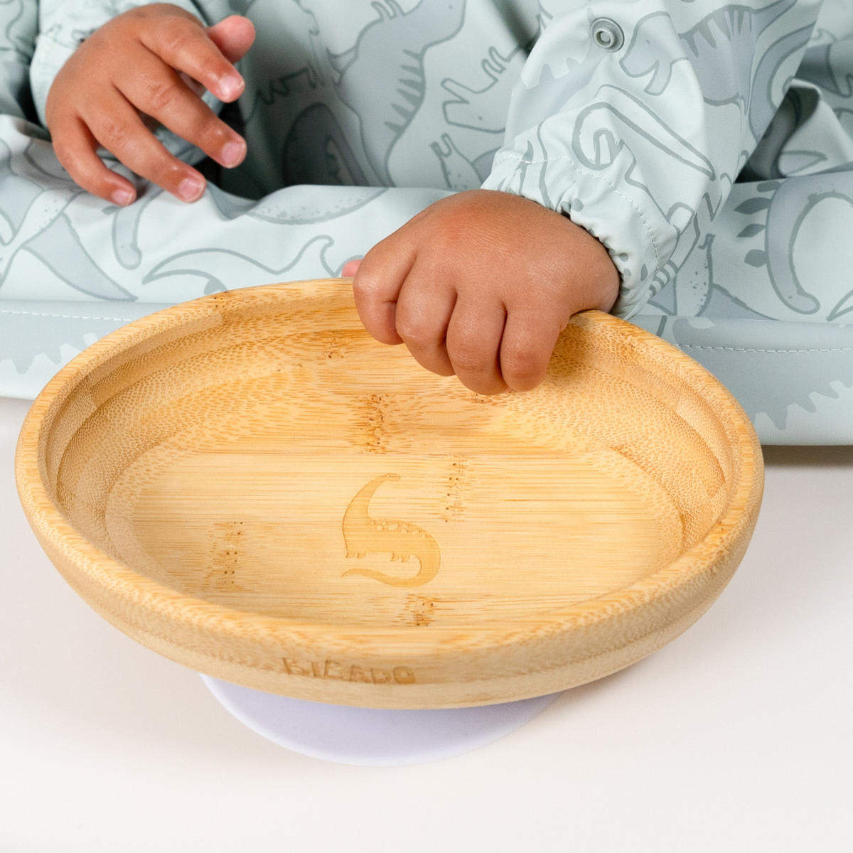 Natural bamboo suction bowl with curved sides and a dinosaur engraving, held by a baby in a mint green bib. - Product image