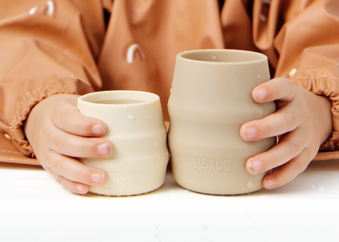 Two beige silicone cups, one smaller than the other, held by a child wearing a brown Coverall bib. The child's hands and torso are visible, and the cups are shown against a clean white background. - Product image