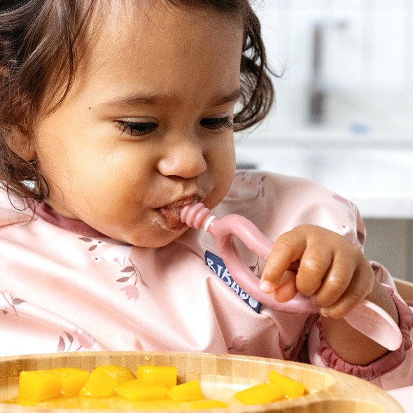 Baby in a pink floral bib uses a pink Dippit™ multi-stage spoon to dip food from a bamboo plate. - Product image