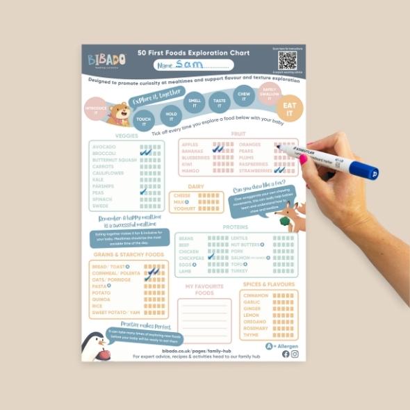 Hand marking a "50 First Foods Exploration Chart" with a blue marker, ticking off strawberries. - Product image