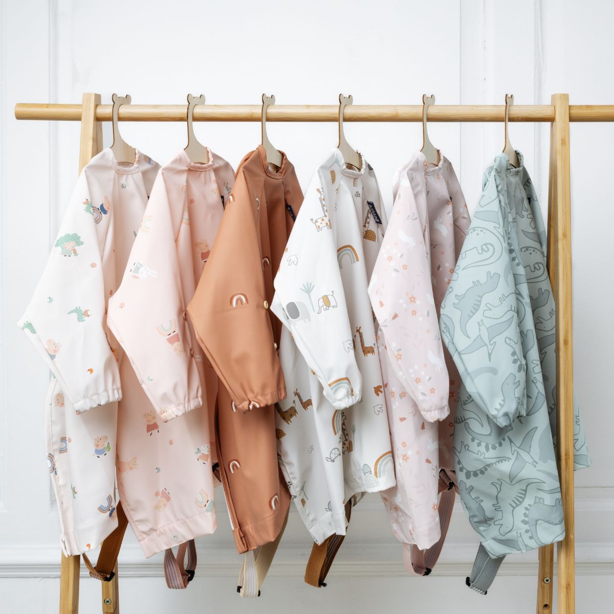 Six long-sleeved baby bibs in various pastel prints like dinosaurs, rainbows, and animals, hanging on a wooden rack. - Product image