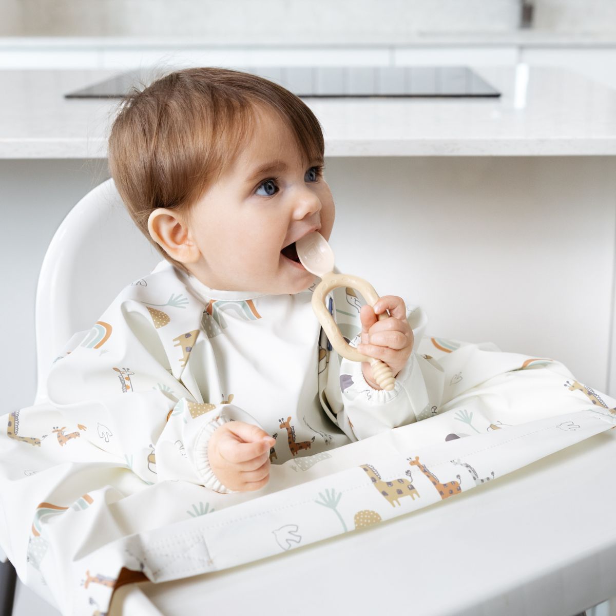 Baby in a white long-sleeve weaning bib with giraffe and rainbow print, holding a wooden teether. - Product image