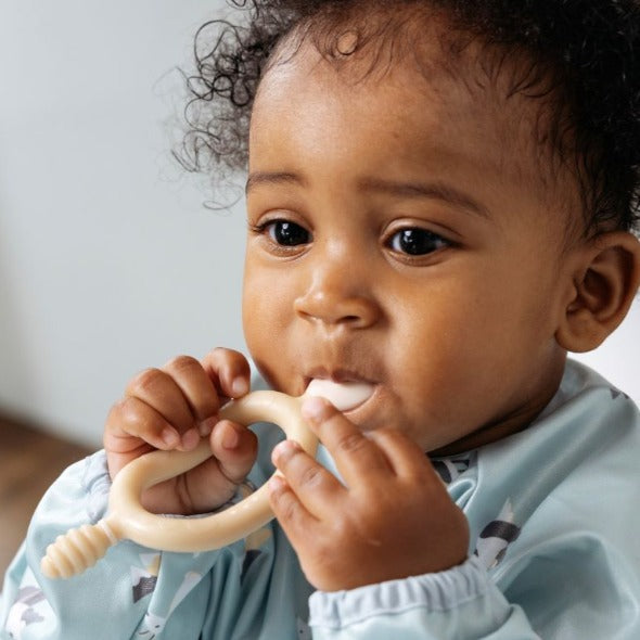 Baby holds a beige silicone Dippit™ dual-ended spoon and textured dipper, wearing a light blue bib. - Product image