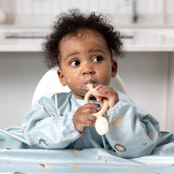 Baby in a light blue long-sleeve weaning bib with animal print, holding a beige Dippit™ or Sporkit™. - Product image