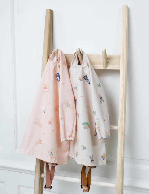 Two long-sleeve coverall weaning bibs, one pink with bunny print and one cream with animal print, hanging on a wooden ladder. - Product image