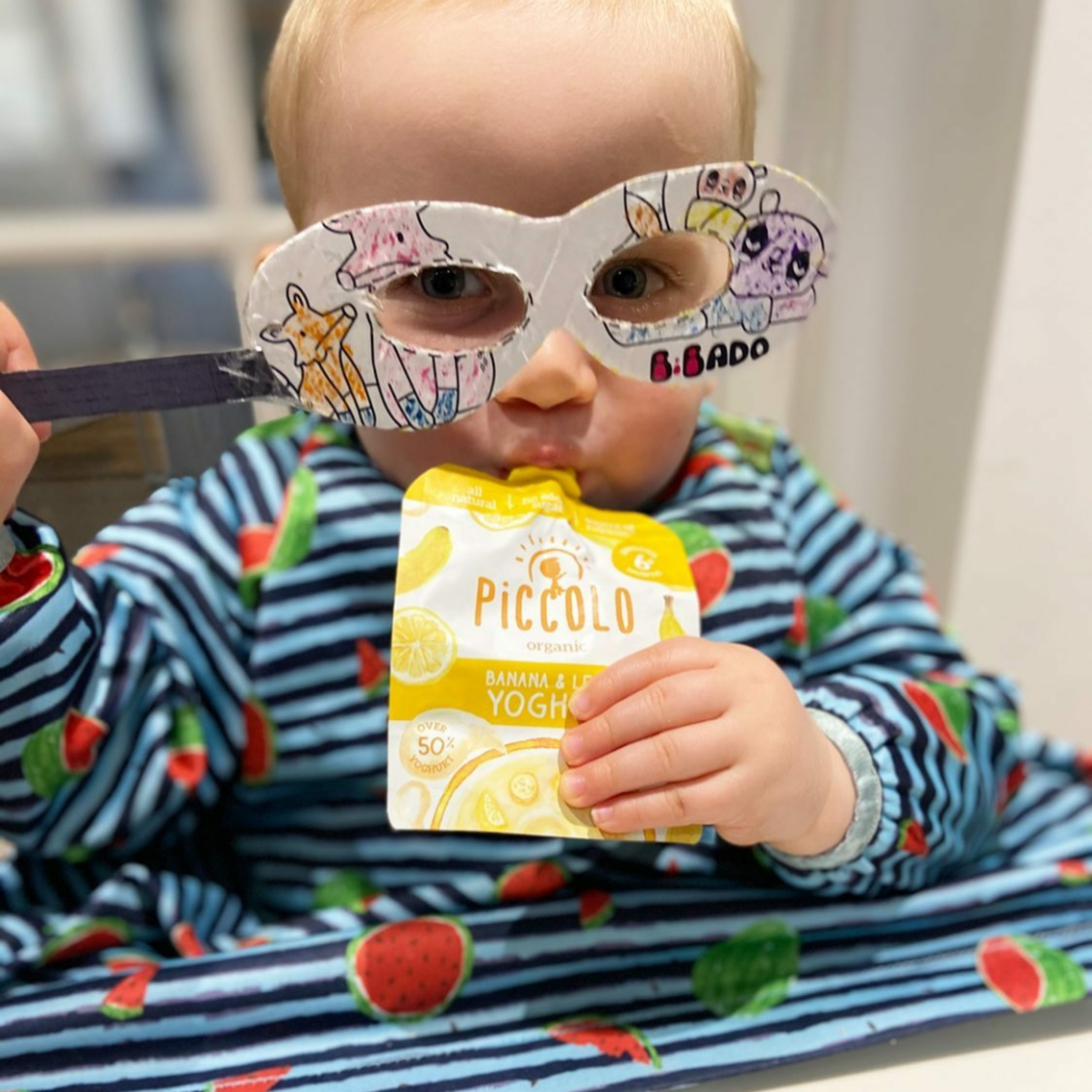 Toddler in a striped bib with watermelons, wearing a paper mask and eating Piccolo banana & lemon yoghurt.