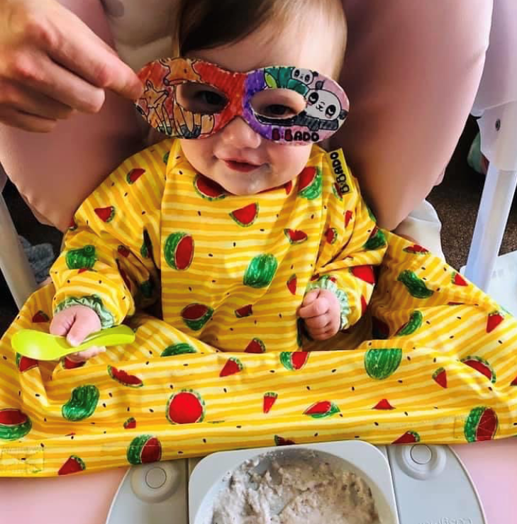 Smiling baby in a yellow watermelon-patterned bib and colourful mask, ready for a meal.