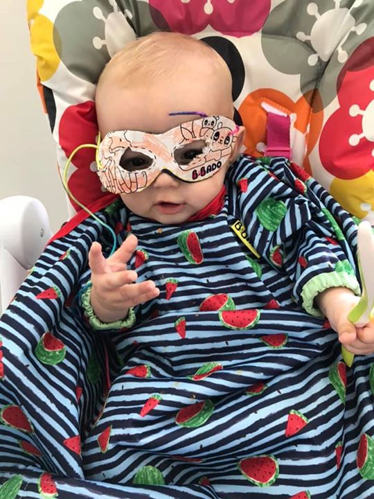 Baby in a highchair wearing a watermelon-print bib and a handmade superhero mask.