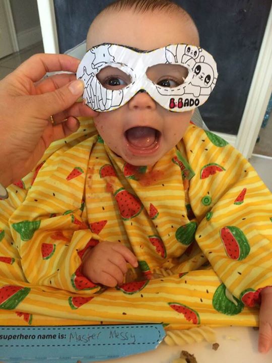 Baby in a yellow watermelon bib, wearing a white superhero mask, mouth open in surprise.