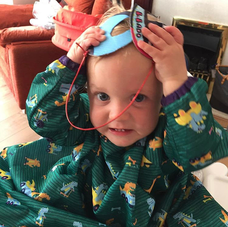 Toddler in a green dinosaur-print cape, holding a blue felt mask with "Bando" written on it.