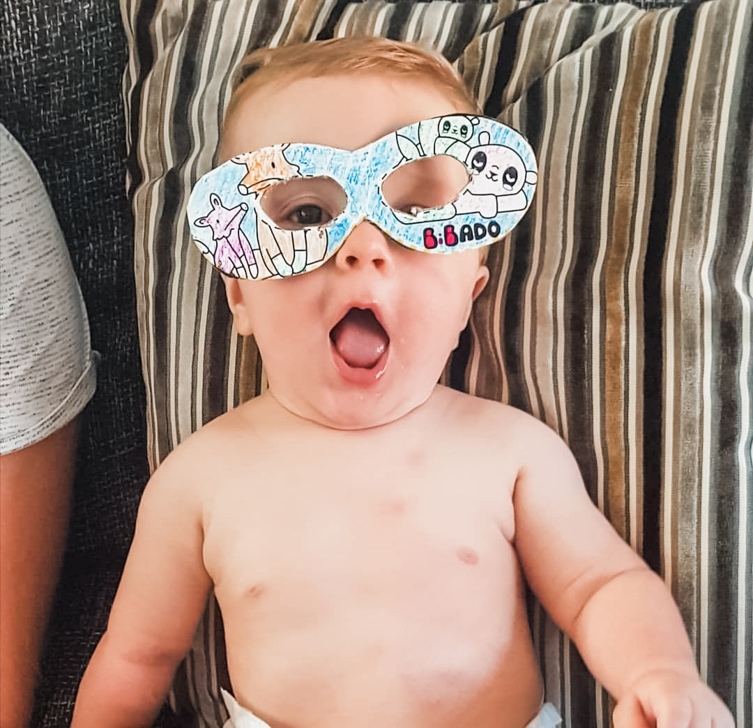 Baby wearing a colourful, hand-drawn paper mask with cartoon animals, lying on a striped cushion.