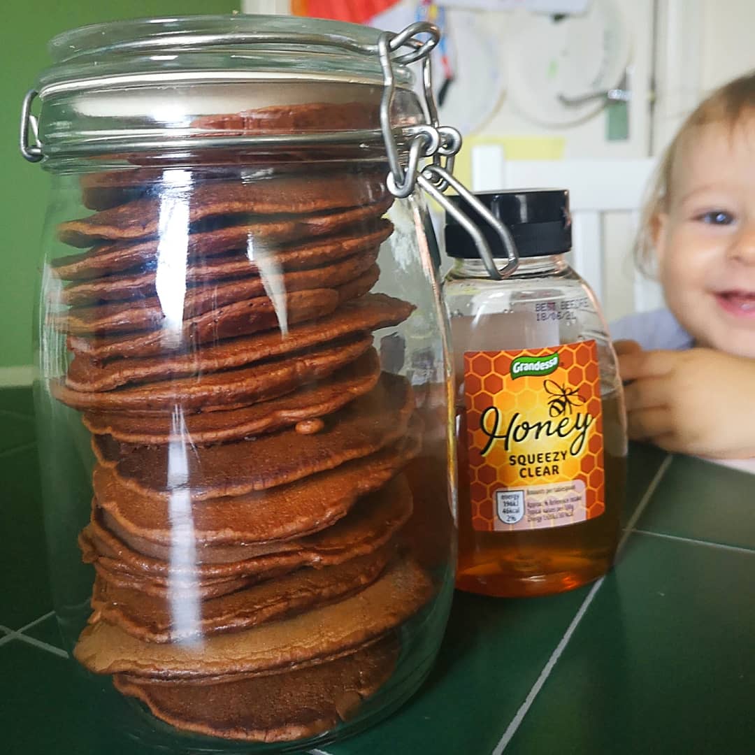 Stack of golden-brown banana cacao pancakes in a glass jar, with honey and a smiling baby.