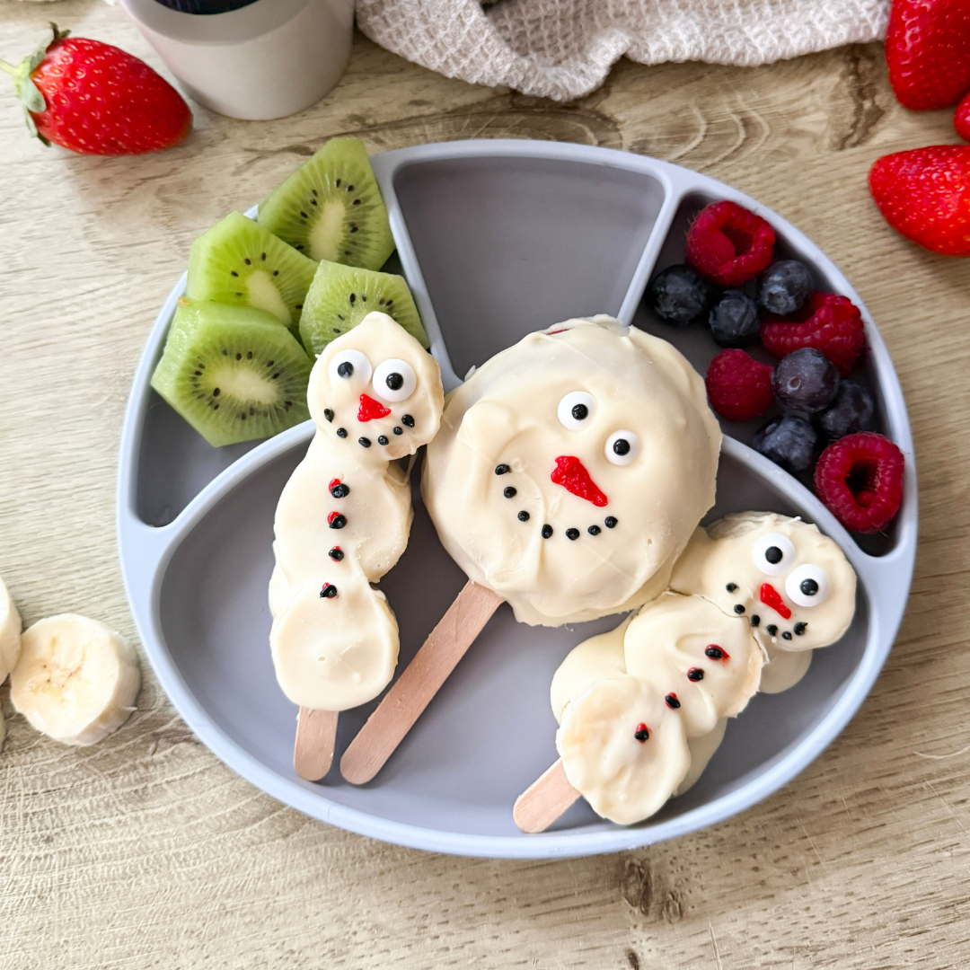 White chocolate snowman apple pops with kiwi, blueberries, and raspberries on a grey plate.