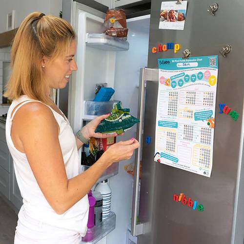 Woman holding a green food pouch, looking at a "50 First Foods Exploration Chart" on a fridge.
