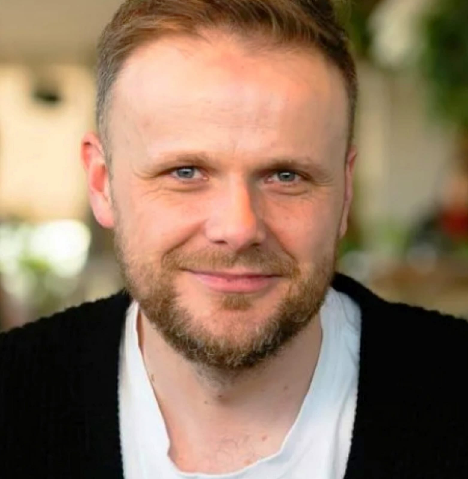 Smiling man with short hair and a neatly trimmed beard, wearing a black cardigan over a white t-shirt.