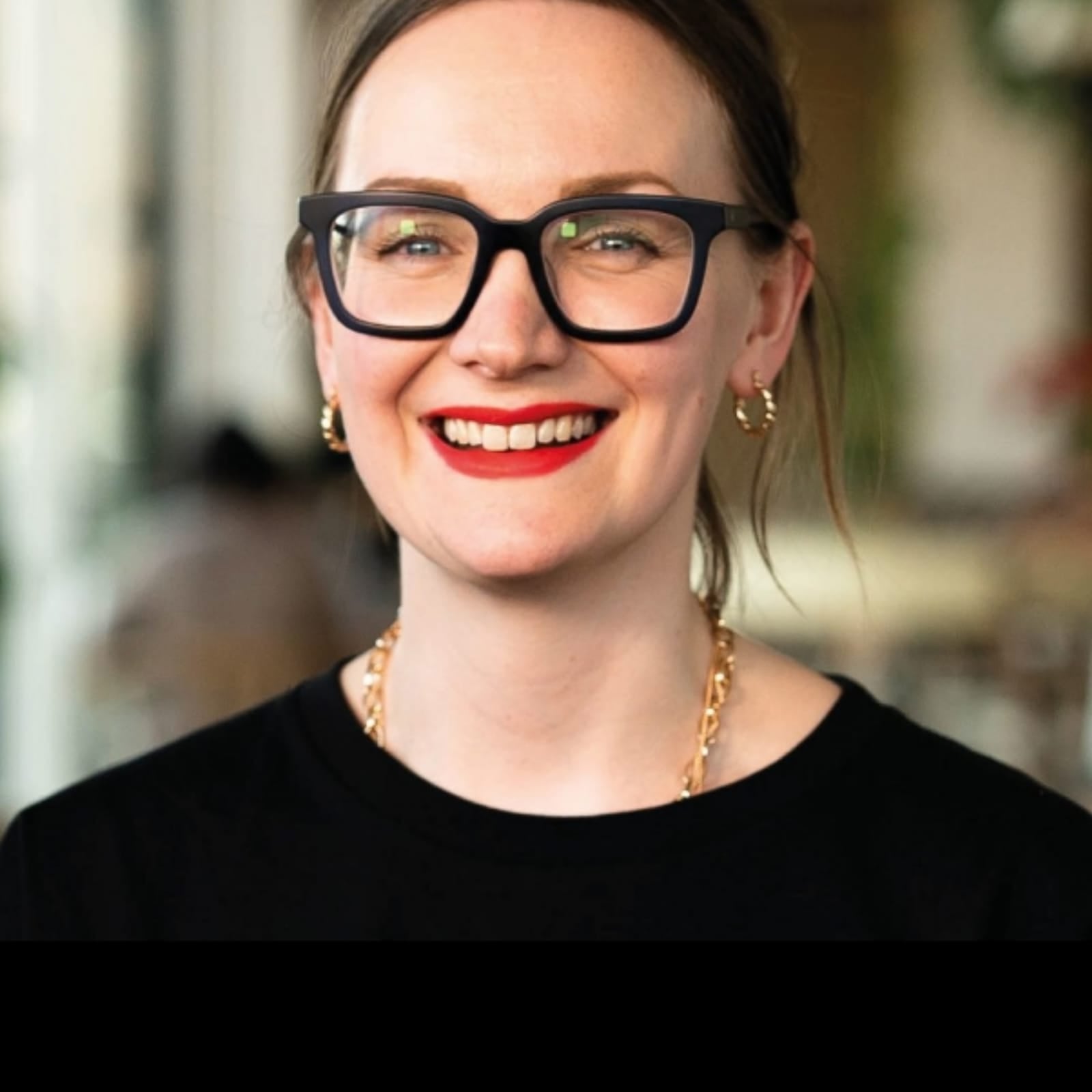 Smiling woman with black glasses, red lipstick, and gold chain necklace.