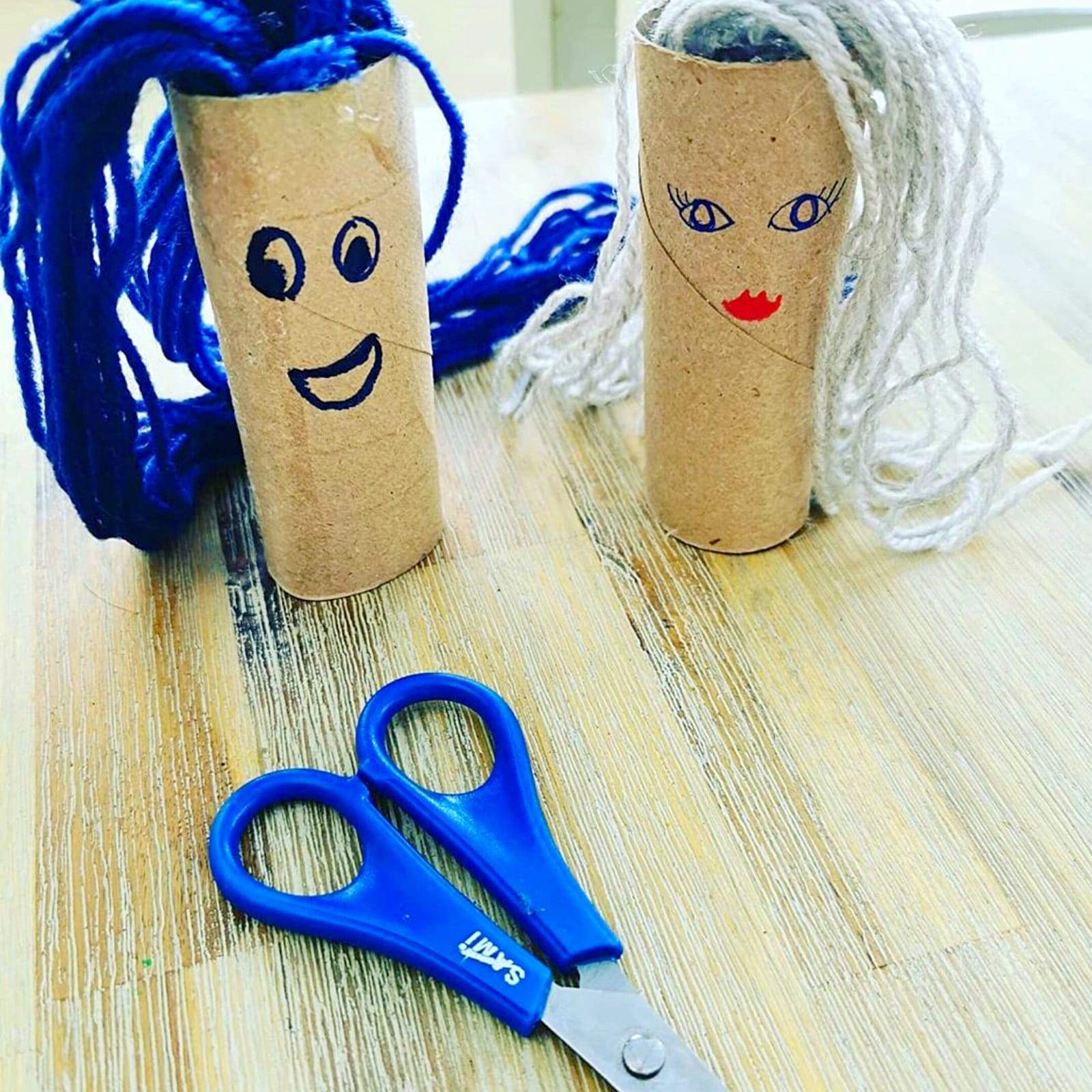 Crafted hairdressing tubes with blue and white yarn hair, alongside blue scissors.