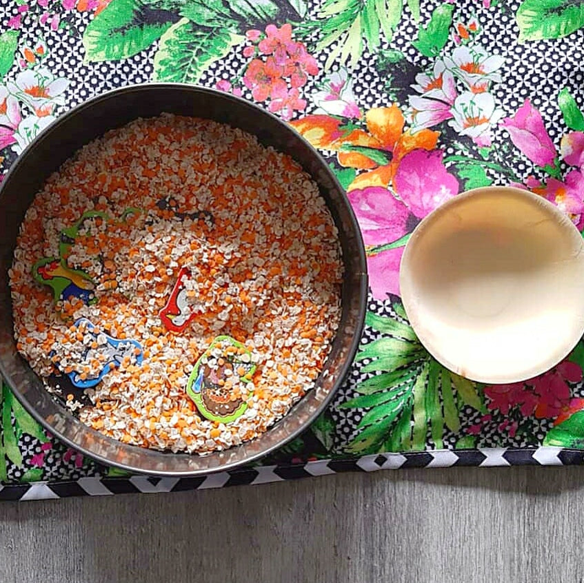 Sensory bin with oats, orange lentils, and colourful animal shapes for a hide 'n' find game.