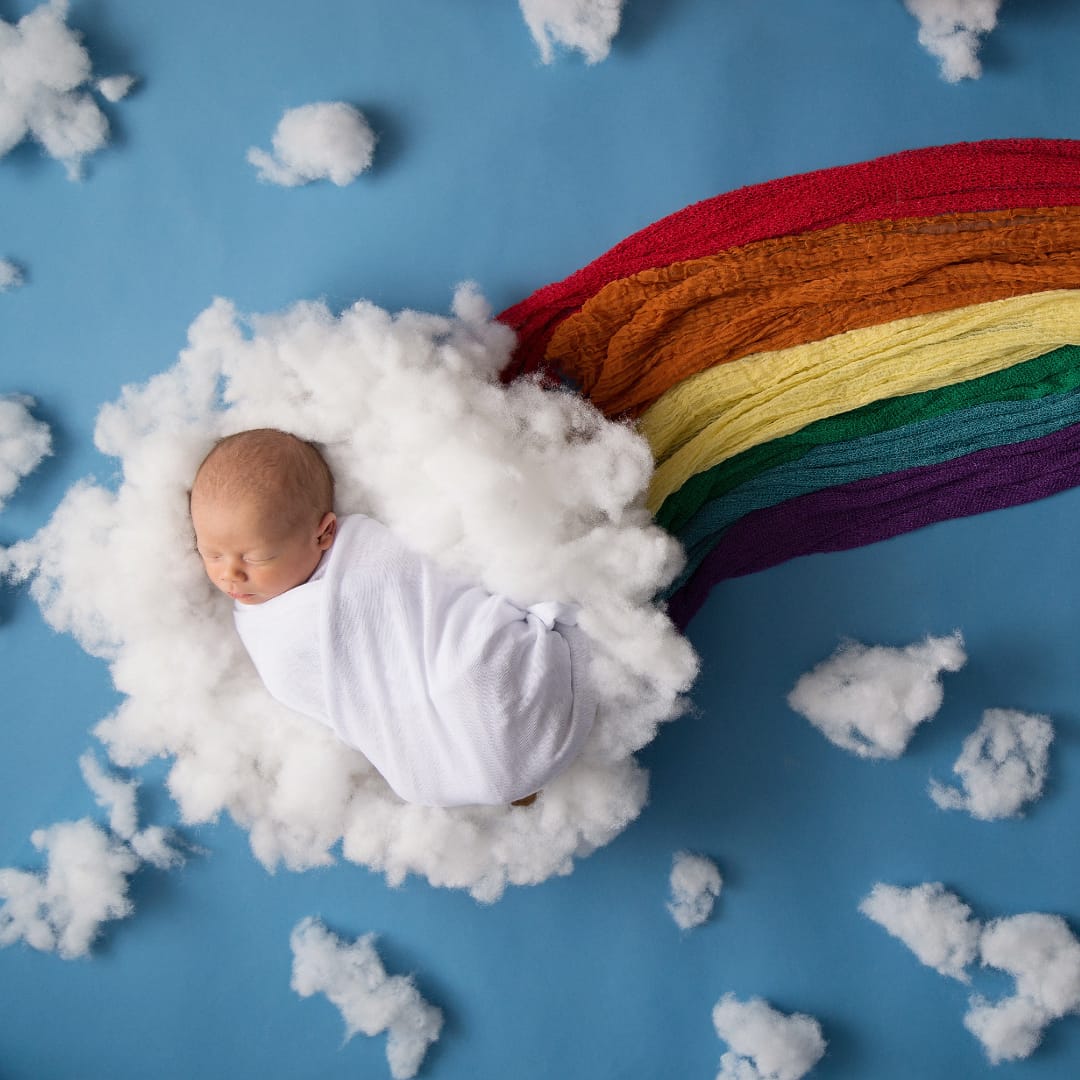 Newborn "rainbow baby" swaddled in white, sleeping on a fluffy cloud with a colourful rainbow.