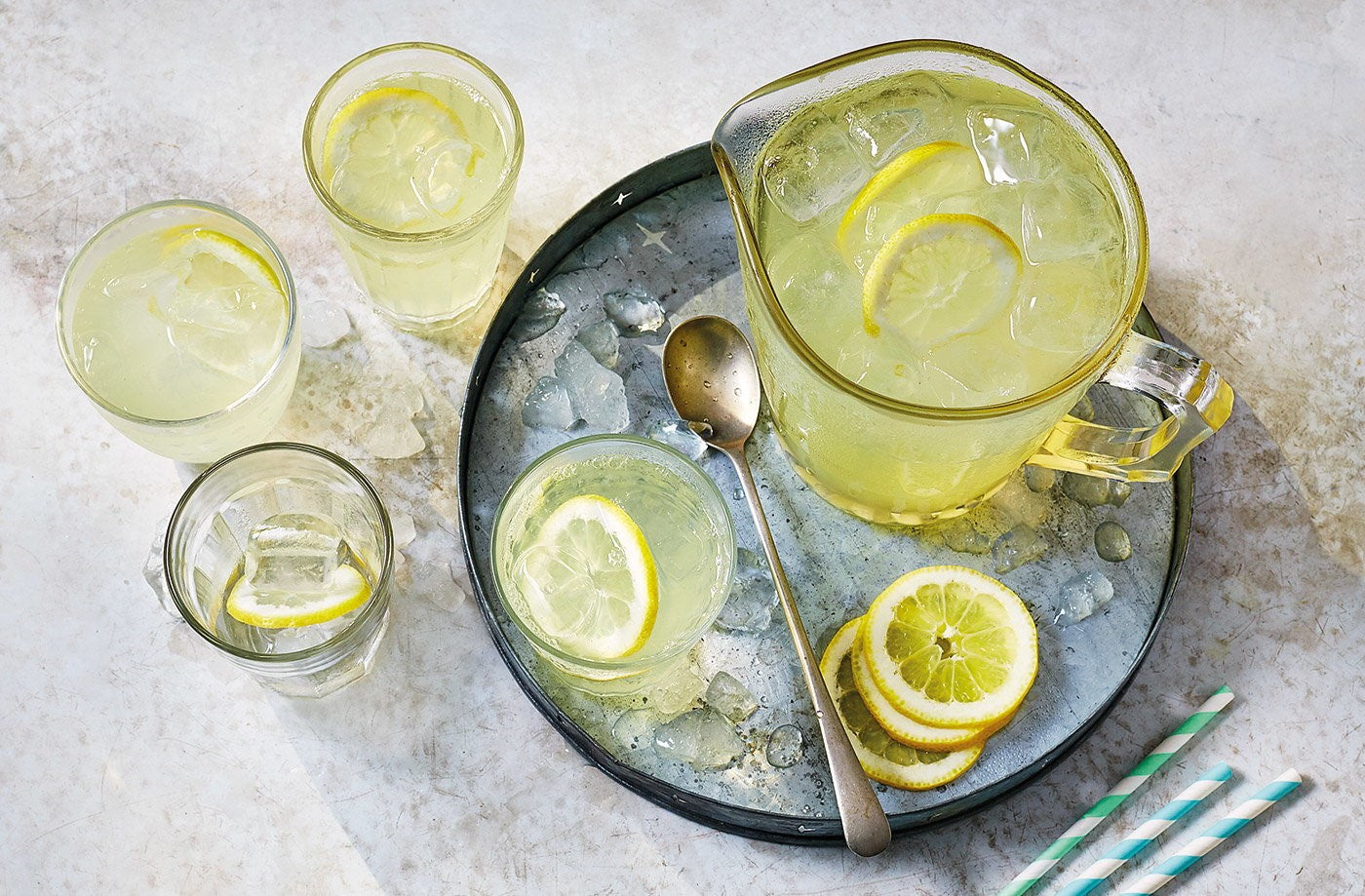 Fresh homemade lemonade in a glass jug and tumblers with lemon slices and ice on a tray.