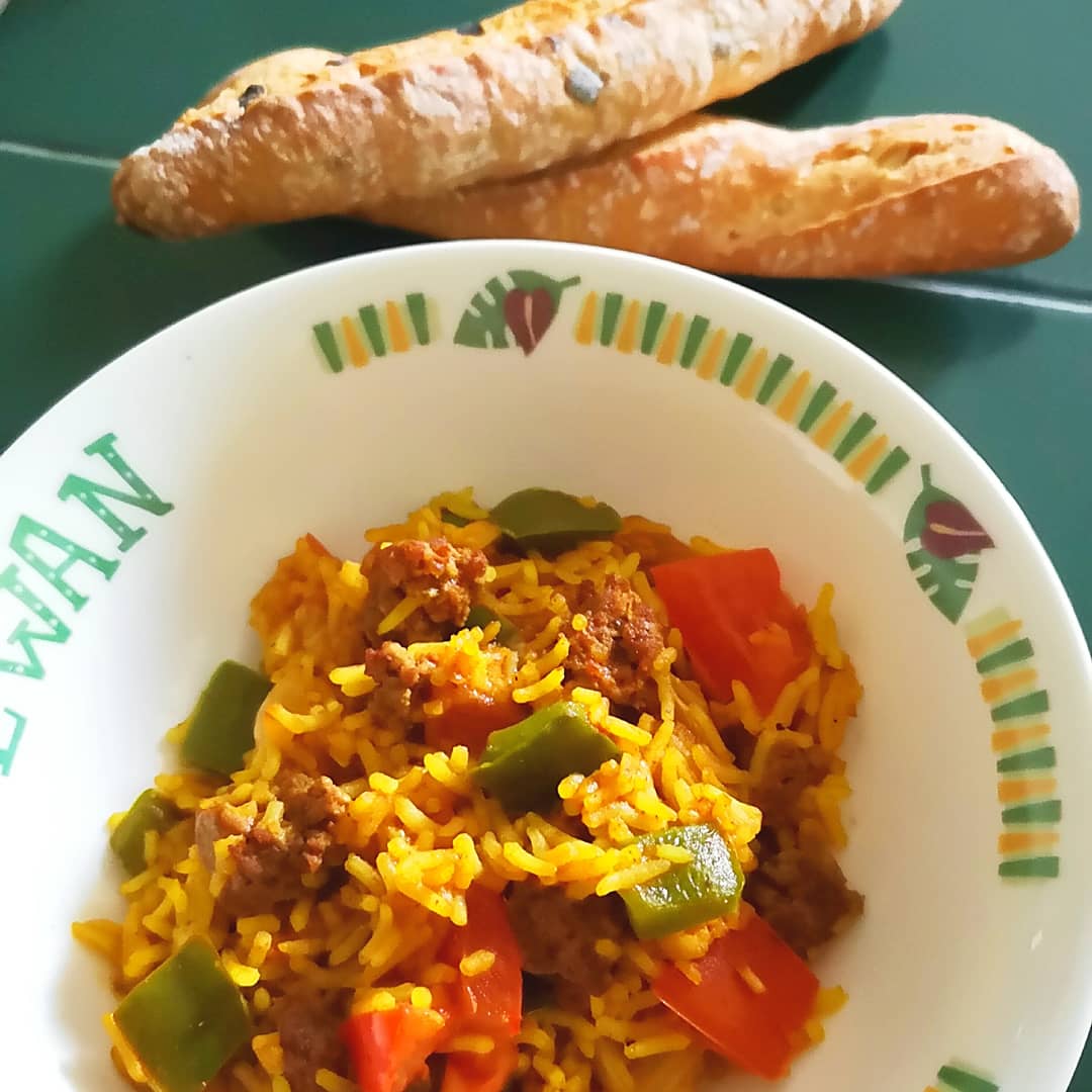 A bowl of vibrant yellow biryani with minced meat, red and green peppers, and two baguettes.