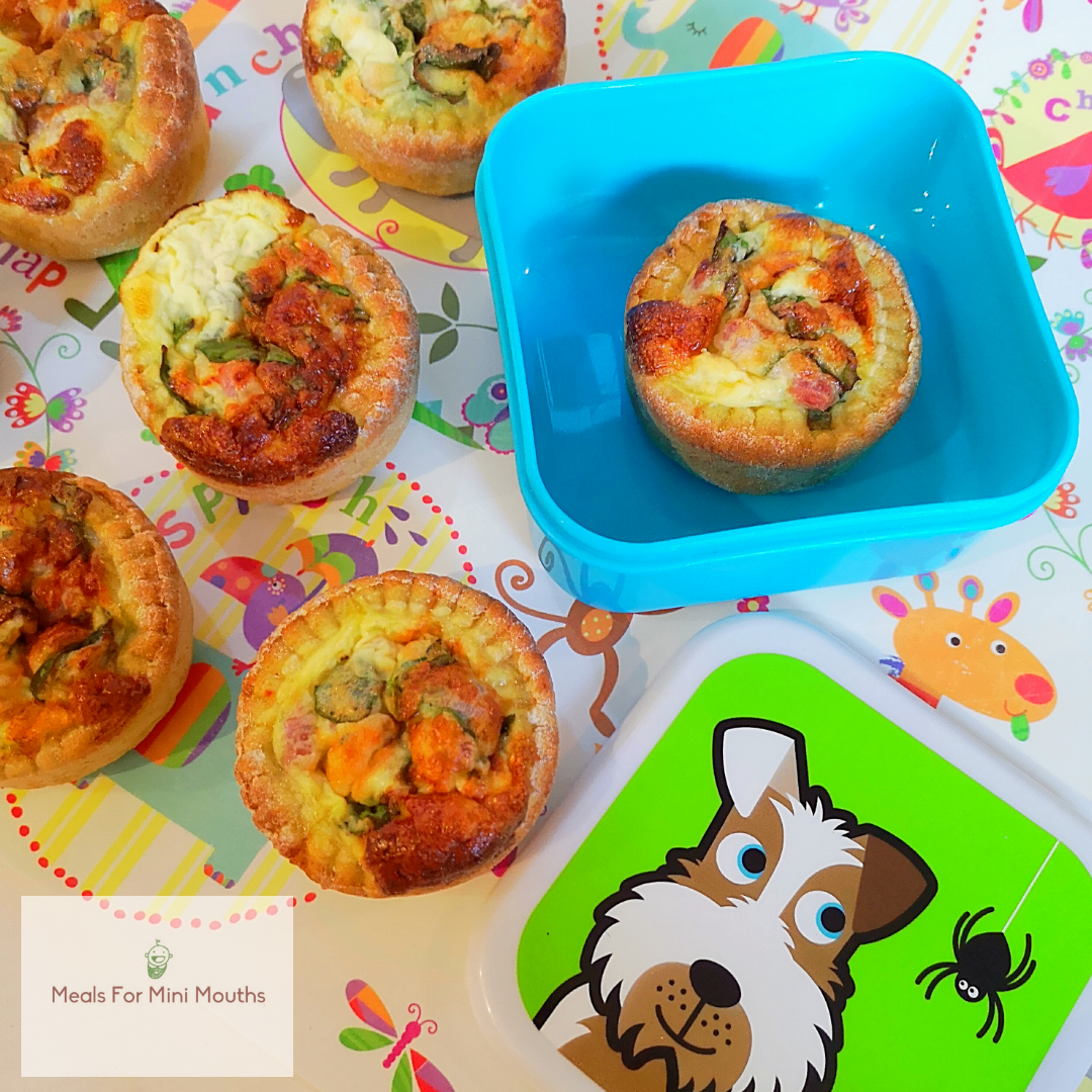 Golden mini quiches, some in a blue lunchbox, on a colourful children's picnic blanket.