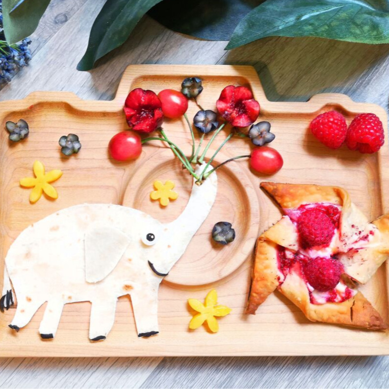 Wooden camera tray with a raspberry pinwheel pastry, tortilla elephant, and fruit flowers.