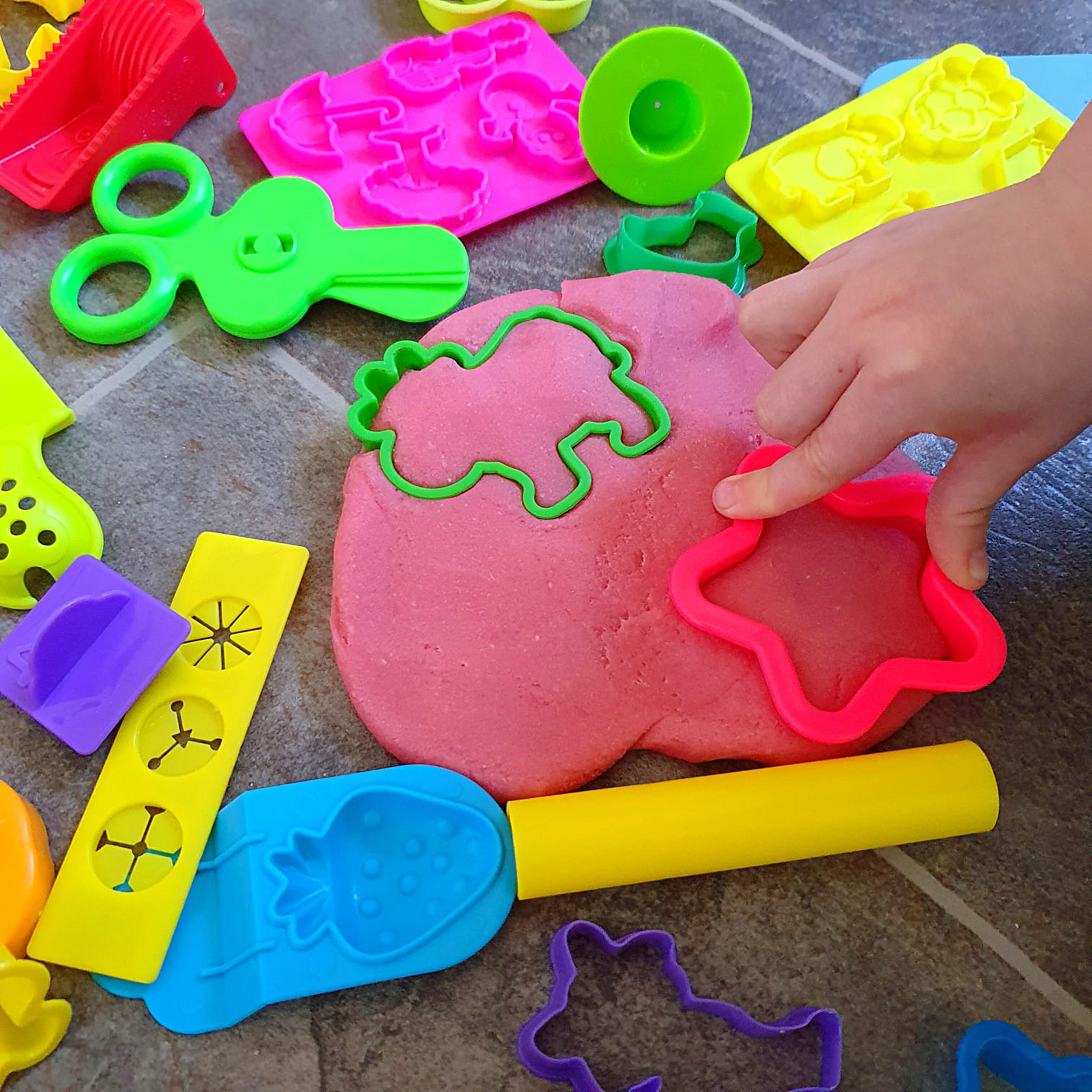 Child's hand using a pink star cutter on homemade pink playdough with colourful tools.