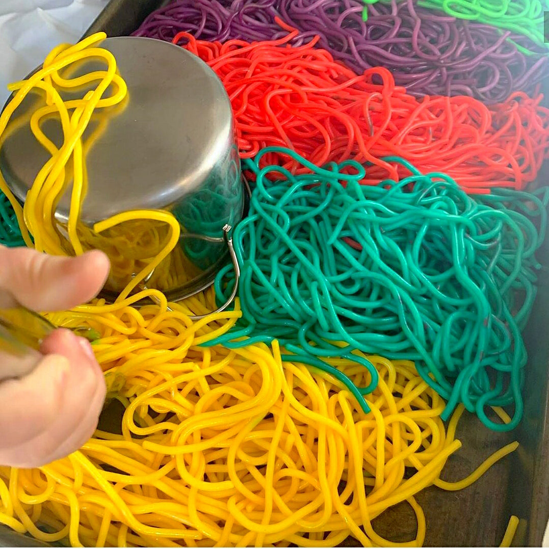 Child's hand playing with vibrant yellow, red, green, and purple spaghetti in a metal tray.