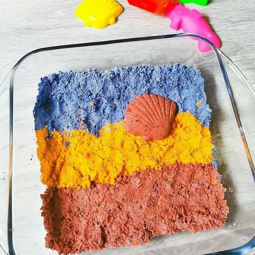 Layered edible sand in blue, yellow, and red with a pink shell, in a glass dish.