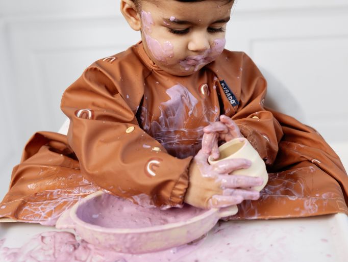 Messy toddler in a brown coverall bib with rainbow print, covered in purple food.