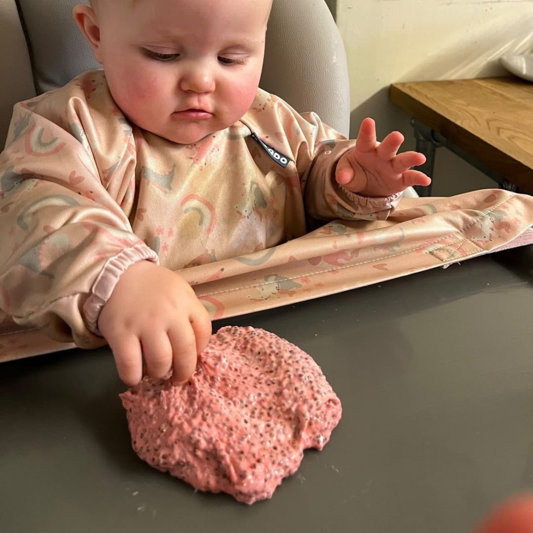Baby in a patterned bib plays with pink chia seed slime on a grey highchair tray.