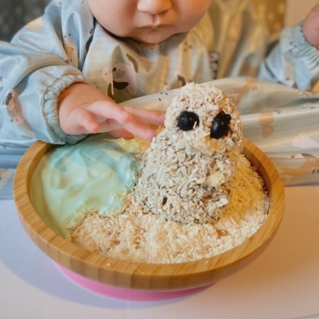 Baby's hand reaching for a wheat biscuit snowman with blueberry eyes in a wooden bowl.